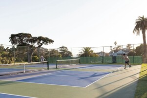 Una cancha de tenis al aire libre con red, rodeada por una valla, árboles y edificios al fondo.