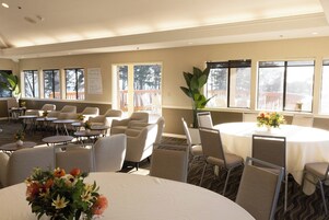 Un salón de conferencias con mesas y sillas redondas, ventanas amplias con vistas, y una pizarra en la pared.