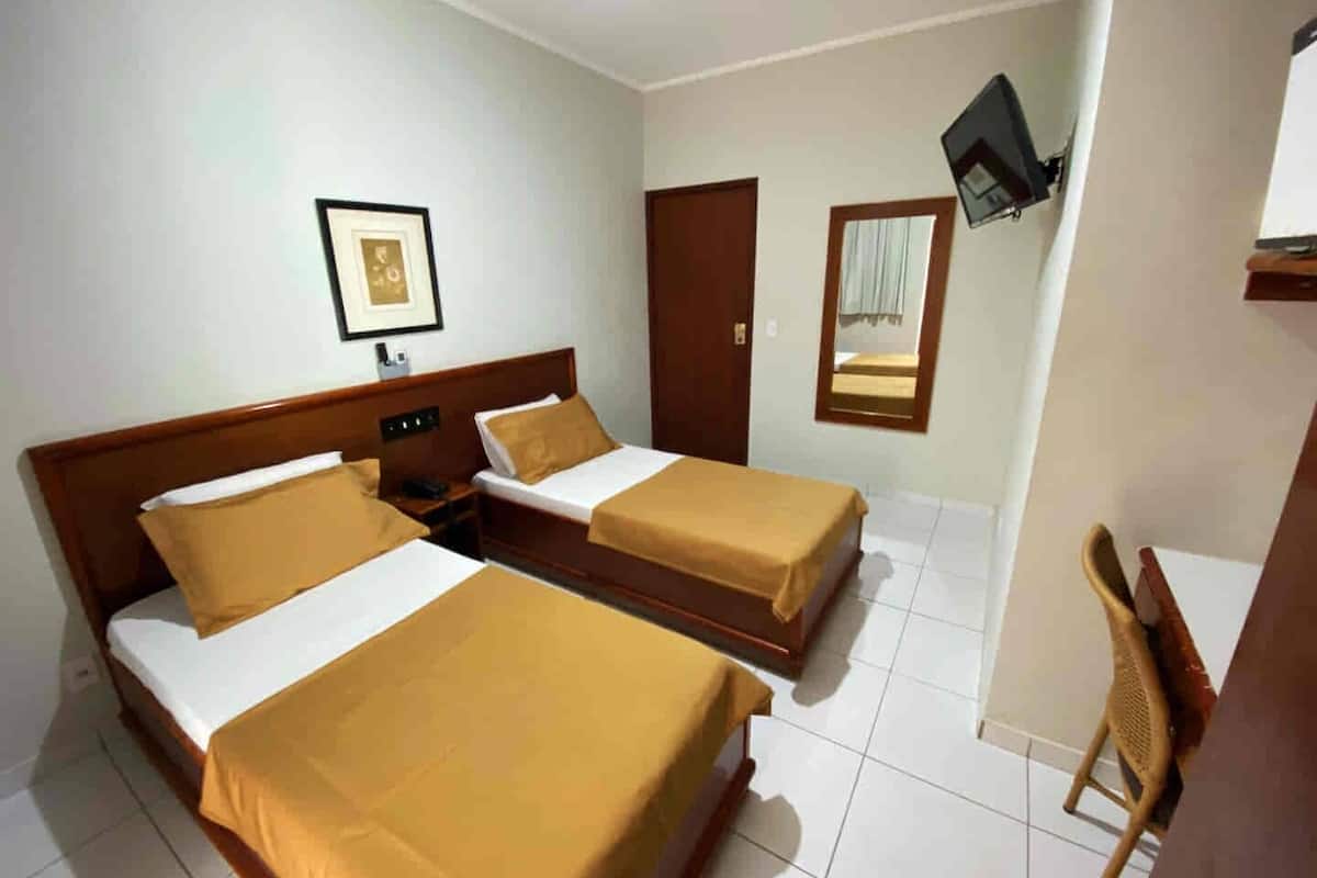 A hotel room with two single beds, a wooden headboard, a small bedside table with a phone, a framed picture on the wall, a mirror, and a TV mounted on the wall.