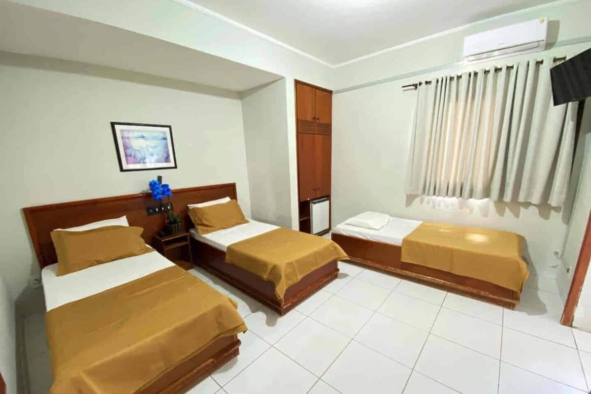 A hotel room with two single beds, a wooden wardrobe, a small desk, a framed picture on the wall, and an air conditioning unit.