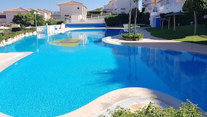 Una piscina amplia y cristalina rodeada de un césped cuidado, con un edificio de balcones y tejado de tejas al fondo.