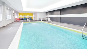 An indoor swimming pool with clear water, surrounded by a modern interior with a yellow accent wall, seating area, and a reception desk.