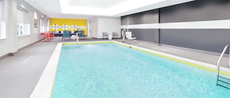 An indoor swimming pool with clear water, surrounded by a modern interior with a yellow accent wall, seating area, and a reception desk.