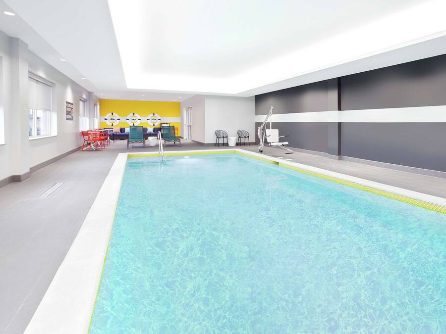 An indoor swimming pool with clear water, surrounded by a modern interior with a yellow accent wall, seating area, and a reception desk.