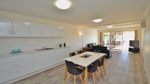 A modern kitchen with white cabinets, a dining area with a white table and black chairs, and a TV in the living area. - Kalbarri Waters A - Kalbarri, WA (Kalbarri)