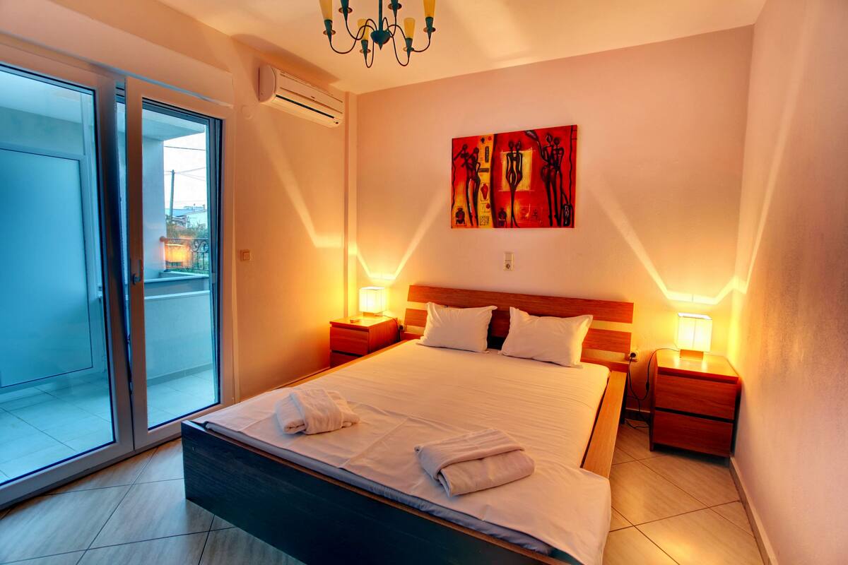 A hotel room with a large bed, two bedside tables, a ceiling light, a wall-mounted artwork, and a sliding glass door leading to an outdoor area.