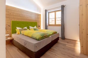 Ein Schlafzimmer mit grauem Matratze und gemusterten Bettwäsche, einem grünen Kopfteil, einem großen Fenster mit Vorhängen und einem Holzboden.