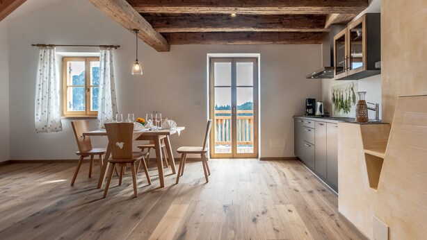 A wooden dining table with chairs, a kitchen with cabinets, and a large window with a view.