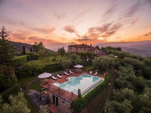 A large rectangular swimming pool surrounded by lounge chairs and umbrellas, with a stone deck and a view of a hilltop building in the background.
