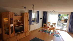 A living room with a wooden entertainment unit, a television, a dining area with a table and chairs, and a large window with curtains. - Vacation cottage Rohde, Germany (Binz)