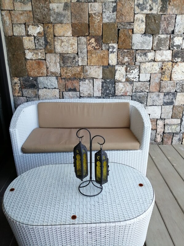 A wicker sofa with a beige cushion against a stone wall, a round wicker table with two black lanterns, and a metal railing.