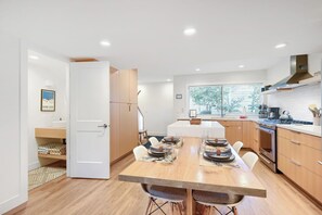 Una cocina moderna con una mesa de comedor para cuatro personas, armarios de madera, horno empotrado y ventana con vista a árboles.