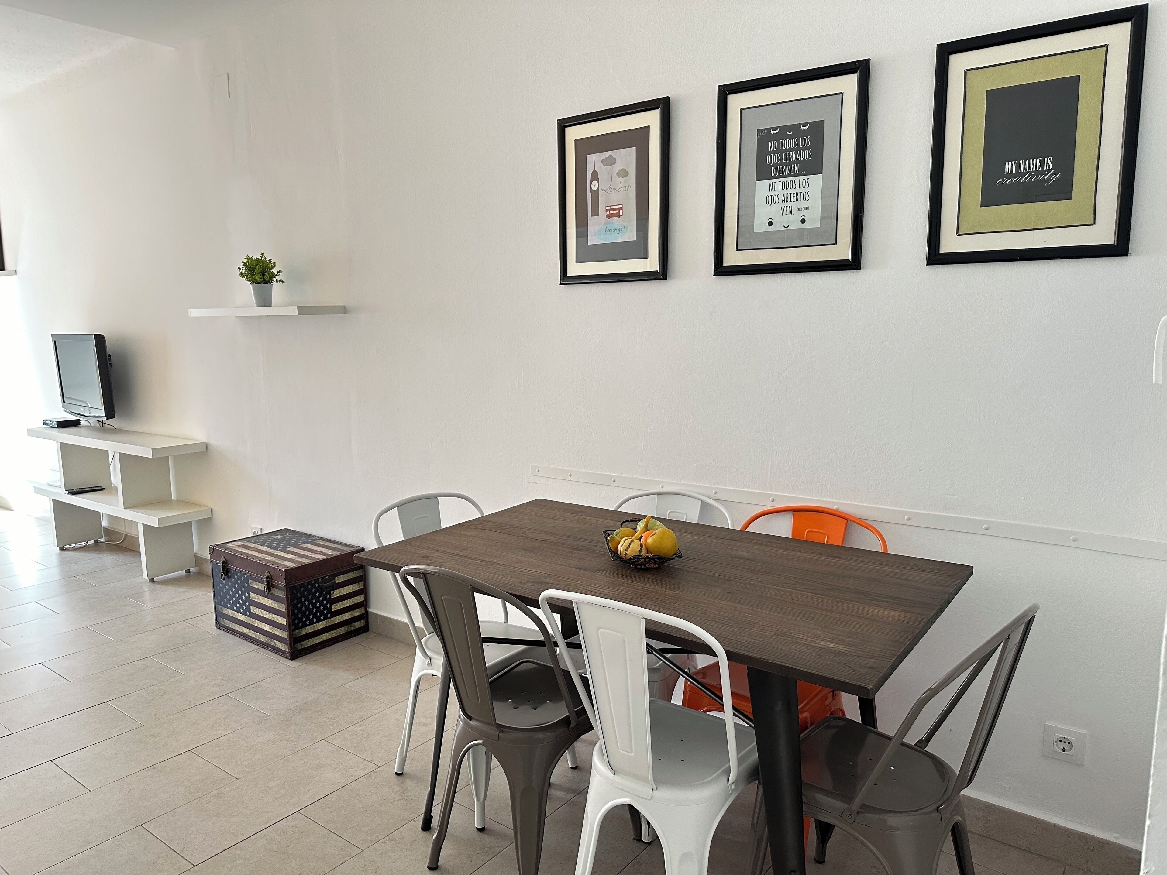 A dining area with a wooden table, white and gray chairs, and a small fruit bowl. There are framed pictures on the wall, a television on a shelf, and a potted plant.