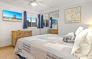 Une chambre avec un grand lit, une commode en bois, un ventilateur de plafond, un tableau représentant des voiliers et une scène de plage sur le mur.