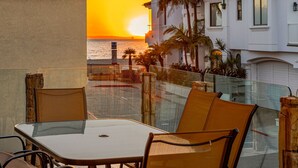 Eine Terrasse mit Glastisch und Stühlen, die auf einen Sonnenuntergang über dem Meer blickt, mit Palmen und einem weißen Gebäude im Hintergrund.