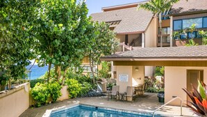 A pool area with a view of the ocean, surrounded by lush greenery and a building with a tiled roof.