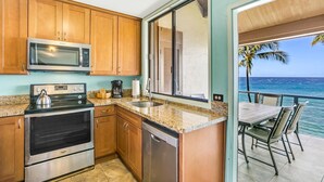 A kitchen with wooden cabinets, a microwave, stove, and a coffee maker. There's a window with a view of the ocean and a patio with chairs.