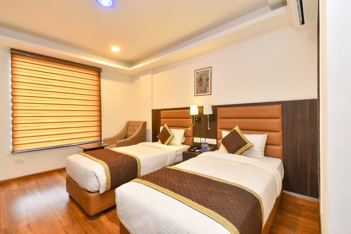 A hotel room with two beds, a wooden floor, a window with blinds, a chair, and a nightstand with a lamp.