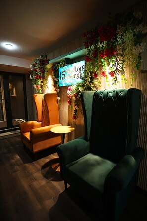 Una habitación con un sillón verde, un sillón naranja, una pequeña mesa redonda y un televisor instalado en la pared que muestra '15th Floor Lounge'.
