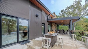 Un patio moderno al aire libre con chimenea de piedra, asientos y un espacio cubierto con vistas a árboles.