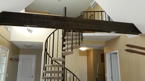 Un escalier en colimaçon, doté d’une main courante en bois foncé et de marches recouvertes d’un tapis beige, mène au deuxième étage, où l’on trouve un ventilateur de plafond et des luminaires encastrés.