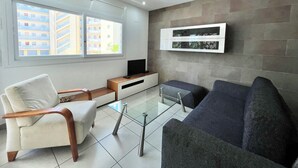Een moderne woonkamer met een bank, fauteuil en glazen salontafel. Een flatscreen-tv is aan de muur bevestigd, en er is een raam met uitzicht op gebouwen.