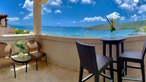 Un balcon agrémenté de fauteuils en osier et d’une petite table, offrant une vue sur une mer turquoise, des bateaux et un paysage vallonné.