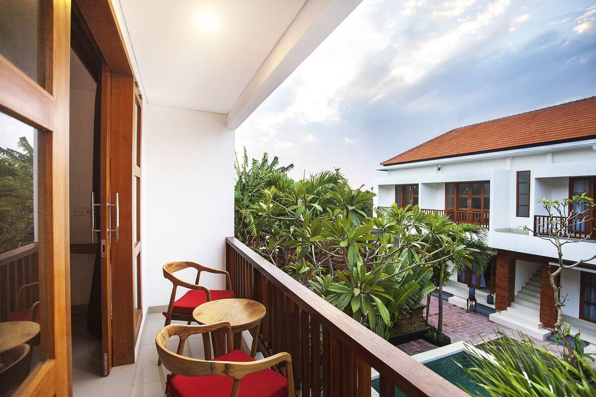 A balcony with wooden chairs and a small table, surrounded by lush greenery and a view of a building with a tiled roof.
