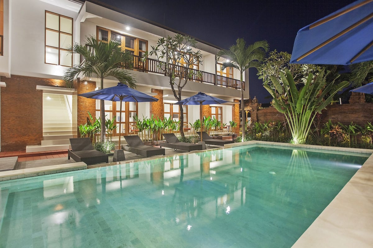 A hotel pool area with lounge chairs and umbrellas, a two-story building with balconies, and a well-maintained garden.