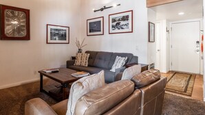 Ein Wohnzimmer mit einer Couch, einem Couchtisch und einem Kamin. An den Wänden hängen Bilderrahmen, und im Hintergrund ist eine Tür zu sehen.
