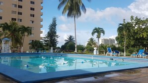 Une piscine entourée de chaises longues et de palmiers, le tout bordé par un immeuble à plusieurs étages.