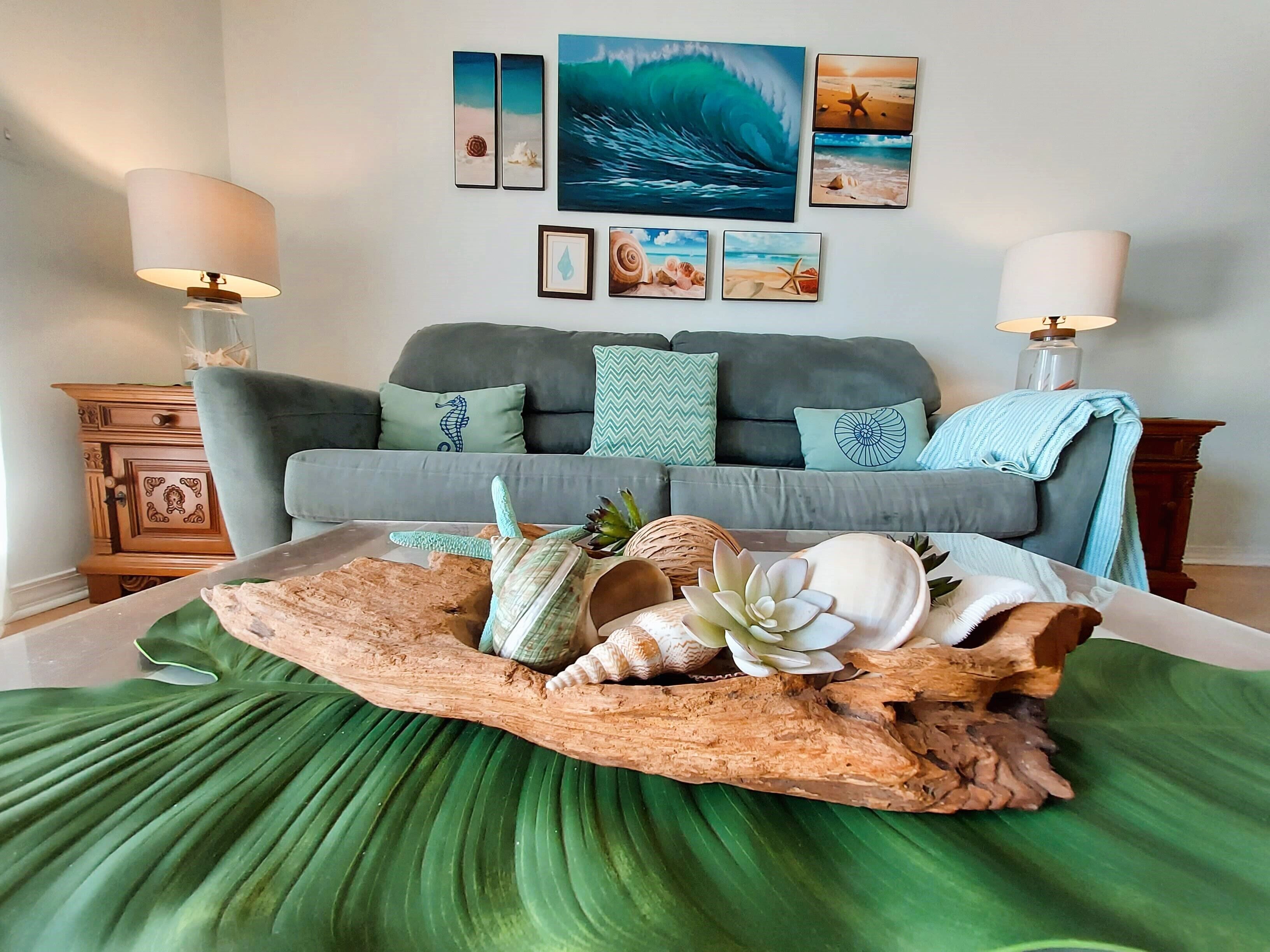 Un salon comprenant un canapé, des coussins décoratifs, une table basse en bois ornée de coquillages et des œuvres d’art murales représentant des scènes marines.