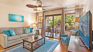A living room with a sofa, coffee table, and TV. There is a ceiling fan, a window with curtains, and a view of the outdoors. - Where Aloha Welcomes You Kihei Condo Private Lanai Pool Steps to the Beach (Kihei)