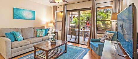 Una sala de estar con sofá, mesa de centro y televisor. Hay un ventilador de techo, una ventana con cortinas y vistas al exterior.