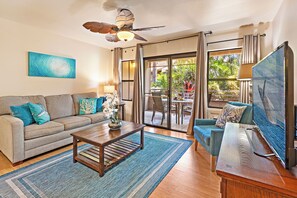 Un salon avec un canapé, une table basse et un téléviseur. On y trouve un ventilateur de plafond, une fenêtre avec des rideaux et une vue sur l’extérieur.