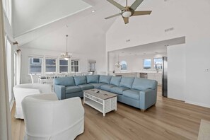 A spacious living room with a sectional sofa, a white armchair, and a wooden coffee table. A ceiling fan is visible, and the room has large windows allowing natural light.