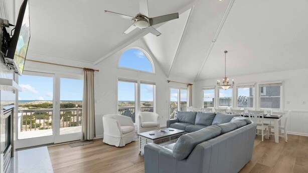 Ein geräumiges Wohnzimmer mit einer großen, grauen Eckcouch, weißen Sessel und einer Essecke mit weißem Tisch und Stühlen. Große Fenster bieten Ausblick auf die Landschaft.