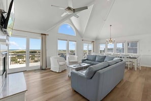 A spacious living room with a large grey sectional sofa, white armchairs, and a dining area with a white table and chairs. There are large windows offering views of the landscape.