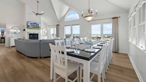 Ein geräumiges Wohnzimmer mit einer großen, grauen Eckcouch, einem Esstisch für sechs Personen und Blick auf den Strand durch große Fenster.