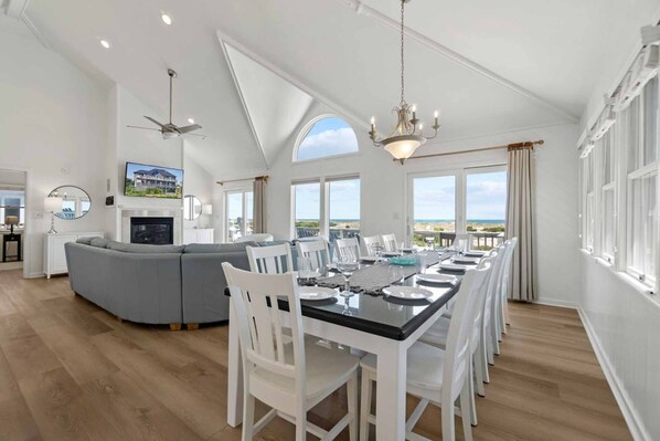 A spacious living room with a large gray sectional sofa, a dining table set for six, and a view of the beach through large windows.