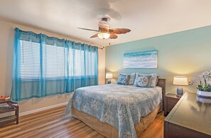 A bedroom with a bed, a ceiling fan, a window with blue curtains, a nightstand, a lamp, and a painting on the wall. - Where Serenity Awaits Kihei Spacious Condo Private Lanai Pool Near Beach (Kihei)