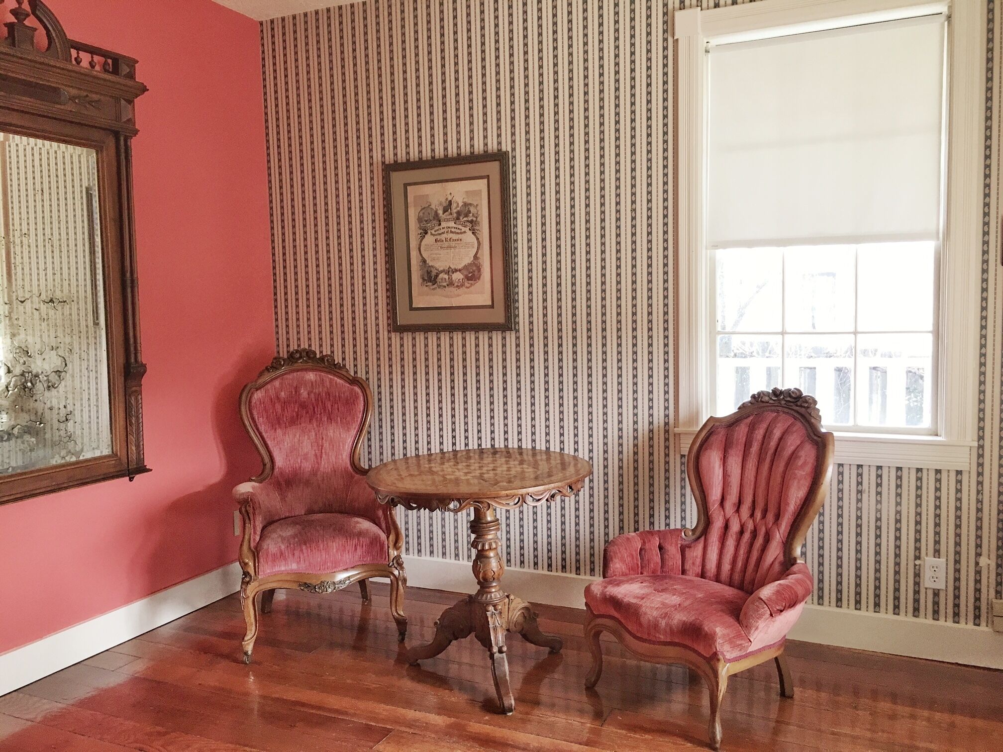 Une pièce avec deux chaises de style vintage, une table ronde en bois et un grand miroir sur le mur de gauche. Les murs sont recouverts d’un papier peint à rayures, et il y a une fenêtre sur le mur de droite.