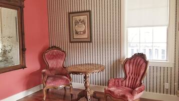 Une pièce avec deux chaises de style vintage, une table ronde en bois et un grand miroir sur le mur de gauche. Les murs sont recouverts d’un papier peint à rayures, et il y a une fenêtre sur le mur de droite.