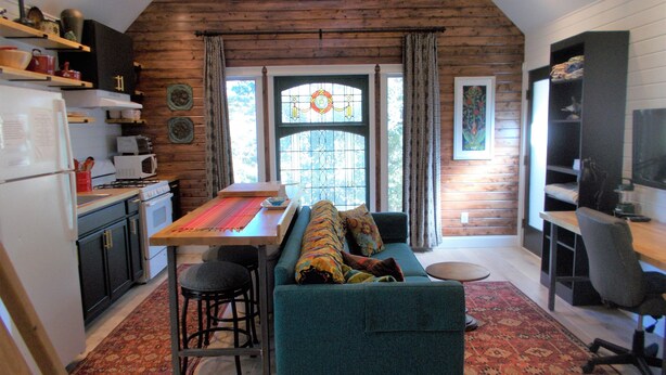 Una cocina acogedora con techo de madera, una mesa de comedor con mantel individual colorido, un sofá verde azulado y un tapete rojo estampado.