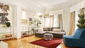 Una sala de estar moderna con piso de madera, un tapete rojo, un sillón azul, un sofá gris y una estantería de vidrio con objetos decorativos.