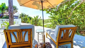 Une terrasse en bois avec deux fauteuils confortables et une table, le tout installé sous un grand parasol, et entouré d’une végétation luxuriante.