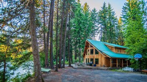 A cabin with a green roof, surrounded by tall trees and a gravel driveway.