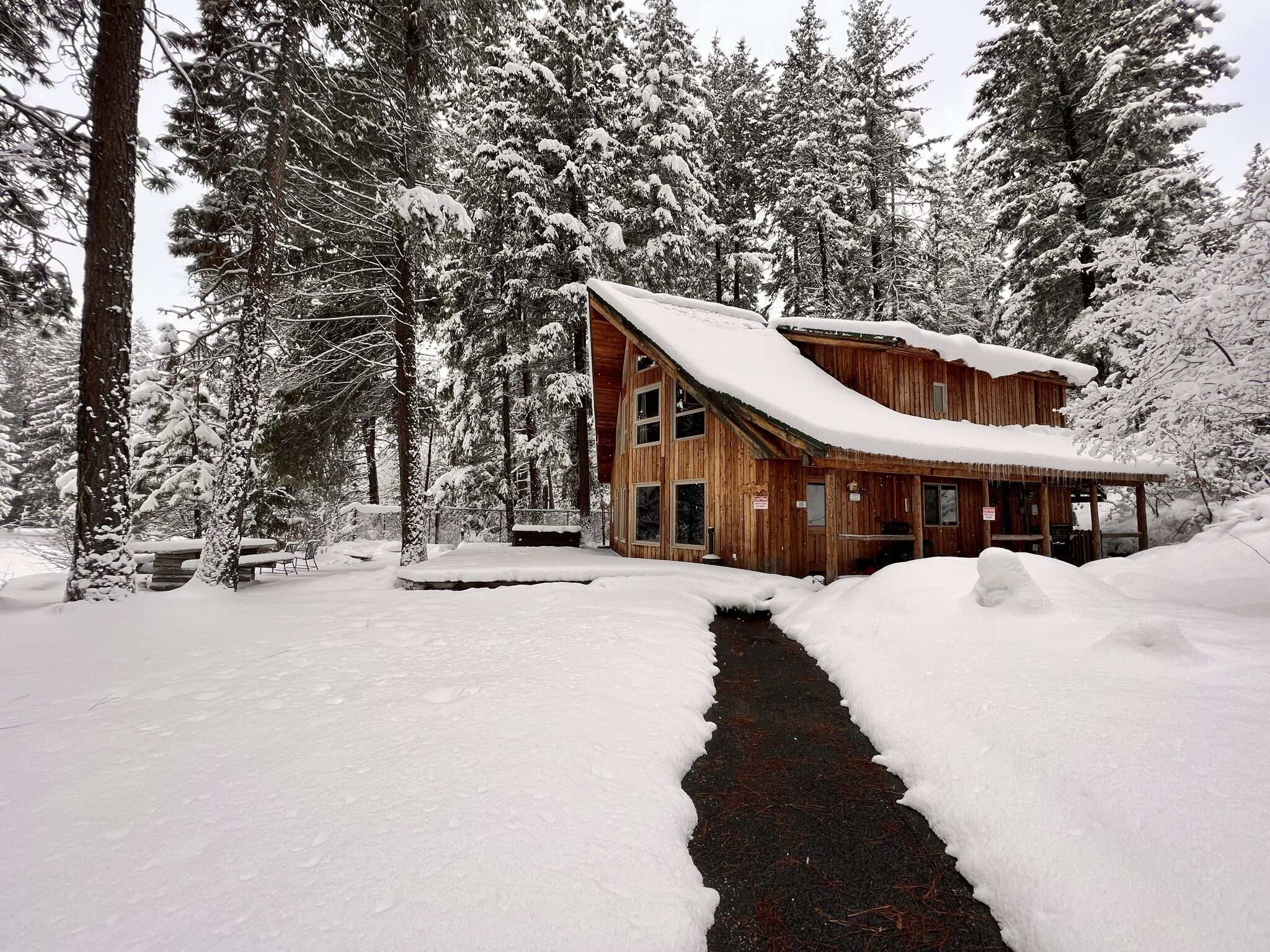Una cabaña de madera con techo nevado y una terraza de madera, rodeada de árboles cubiertos de nieve.