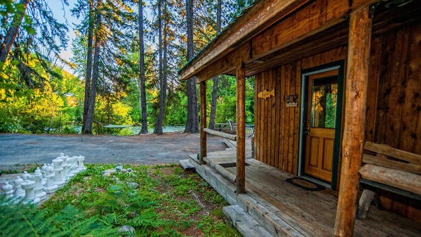 A rustic cabin with a porch, surrounded by tall trees and a gravel path.