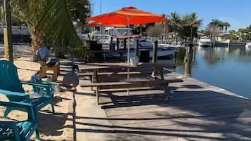 A wooden dock with a picnic table and chairs, a red umbrella, and boats docked in the background.